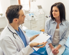 Imagem de um médico segurando uma prancheta e uma caneta e conversando com uma paciente sentada na maca